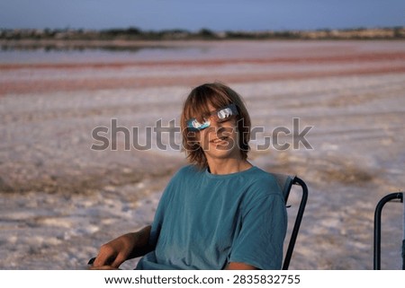 Young boy wearing solar eclipse glasses, sitting in a camping chair and looking at the camera, enjoying the view of the vast pink salt lake and its unique colors during a calm evening