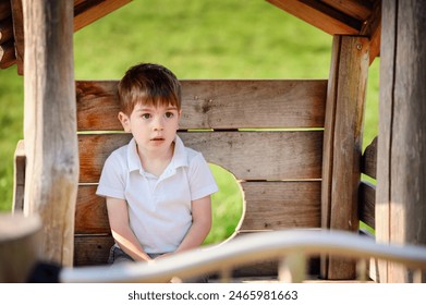 A young boy in a white shirt sits inside a wooden playhouse at the park, looking thoughtful as he enjoys a sunny day outdoors. - Powered by Shutterstock