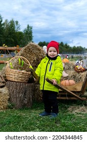 Young Boy With Wooden Cart In His Hand. Little Boy In Bright Green Jacket And Red Hat With Fall Props Around. Autumn Open Air And Festival.