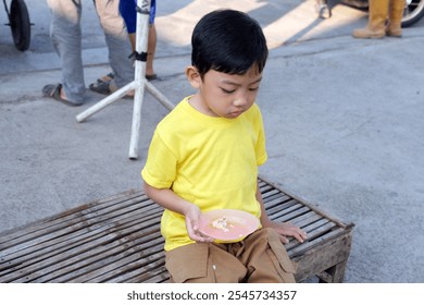  A young boy in a yellow shirt holds a plate of food outdoors, appearing thoughtful. Perfect for content focused on children, outdoor activities, or traditional food and local culture. - Powered by Shutterstock