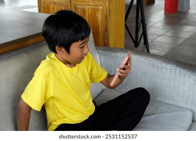 A young boy in a yellow shirt sits on a couch, engrossed in his phone. The bright natural lighting adds warmth to the scene as he focuses on the screen, possibly engaged in a video call or game. - Powered by Shutterstock