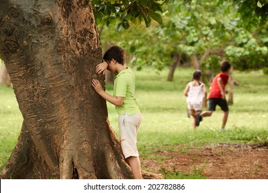 young boys and girls playing hide and seek in park, with kid counting leaning on tree