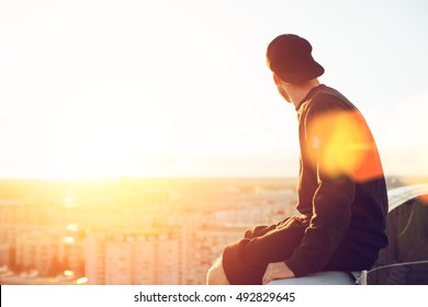 Young and brave man sitting on the edge of the roof and looking far away at the city, lens flares