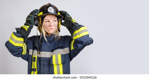 young brave woman in uniform of firefighter puts hardhat on her head and looking away on gray background, copy space for text