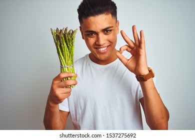 Young Brazilian Man Eating Asparagus Standing Over Isolated Grey Background Doing Ok Sign With Fingers, Excellent Symbol