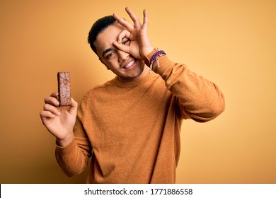 Young brazilian man eating healthy energy bar with protein over isolated yellow background with happy face smiling doing ok sign with hand on eye looking through fingers