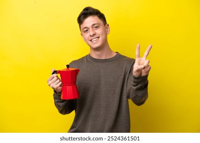 Young Brazilian man holding coffee pot isolated on yellow background smiling and showing victory sign - Powered by Shutterstock