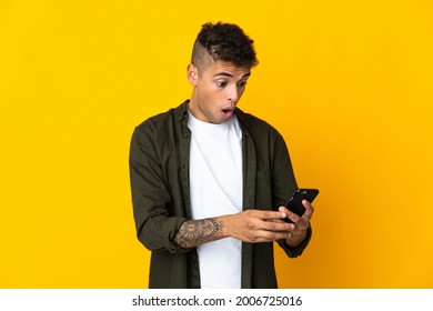 Young brazilian man isolated on yellow background looking at the camera while using the mobile with surprised expression