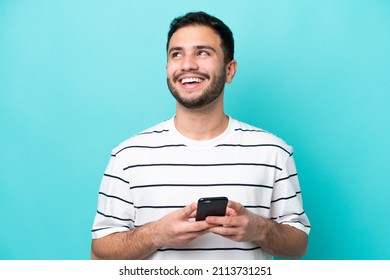 Young Brazilian man isolated on blue background using mobile phone and looking up - Shutterstock ID 2113731251