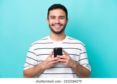 Young Brazilian man isolated on blue background looking at the camera and smiling while using the mobile