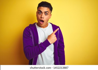 Young brazilian man wearing purple sweatshirt standing over isolated yellow background Surprised pointing with finger to the side, open mouth amazed expression.