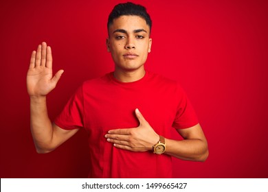 Young brazilian man wearing t-shirt standing over isolated red background Swearing with hand on chest and open palm, making a loyalty promise oath