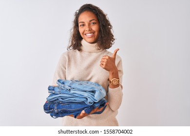 Young Brazilian Shopkeeper Woman Holding Pile Of Jeans Over Isolated White Background Happy With Big Smile Doing Ok Sign, Thumb Up With Fingers, Excellent Sign