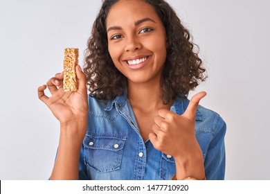 Young Brazilian Woman Eating Granola Bar Standing Over Isolated White Background Happy With Big Smile Doing Ok Sign, Thumb Up With Fingers, Excellent Sign