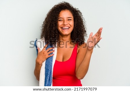 Young brazilian woman going to the beach holding a towel isolated on blue background receiving a pleasant surprise, excited and raising hands.