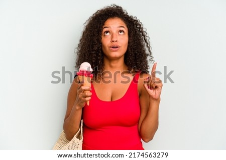 Young brazilian woman going to the beach holding an ice cream isolated on blue background pointing upside with opened mouth.