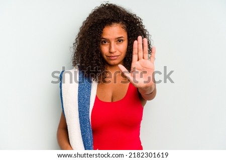 Young brazilian woman going to the beach holding a towel isolated on blue background standing with outstretched hand showing stop sign, preventing you.