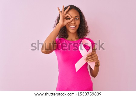 Young brazilian woman holding cancer ribbon standing over isolated pink background with happy face smiling doing ok sign with hand on eye looking through fingers