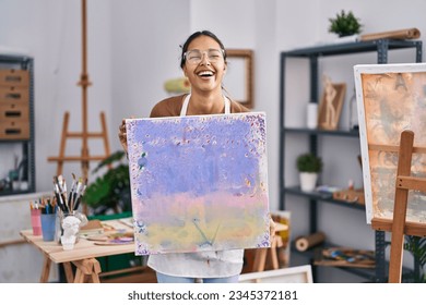 Young brazilian woman holding painter canvas at art studio smiling and laughing hard out loud because funny crazy joke. 