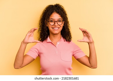 Young Brazilian Woman Isolated On Yellow Background Holding Something With Palms, Offering To Camera.