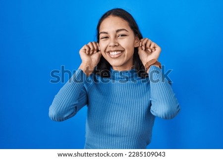 Young brazilian woman standing over blue isolated background smiling pulling ears with fingers, funny gesture. audition problem 