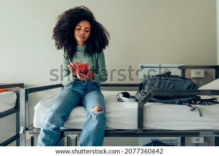 young brazilian woman with thick curly hair sitting on hostel bunk bed smiles using phone