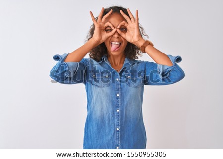 Young brazilian woman wearing denim shirt standing over isolated white background doing ok gesture like binoculars sticking tongue out, eyes looking through fingers. Crazy expression.