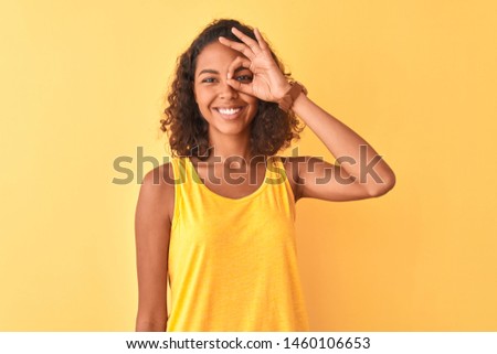 Young brazilian woman wearing t-shirt standing over isolated yellow background doing ok gesture with hand smiling, eye looking through fingers with happy face.