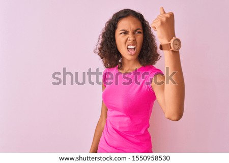 Young brazilian woman wearing t-shirt standing over isolated pink background angry and mad raising fist frustrated and furious while shouting with anger. Rage and aggressive concept.