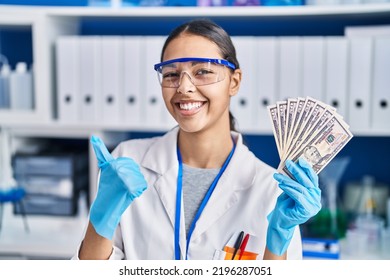 Young Brazilian Woman Working At Scientist Laboratory Holding Money Smiling Happy And Positive, Thumb Up Doing Excellent And Approval Sign 