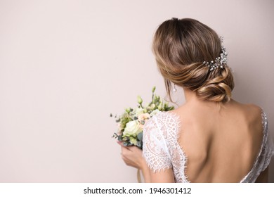 Young bride with elegant hairstyle holding wedding bouquet on beige background, back view. Space for text