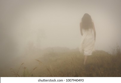 Young bride with long hair in long white wedding dress going in foggy mist landscape. Blur discernible outline of slim figure in fog on left side of photo. View from side. Mistery landscape - Shutterstock ID 1007940493