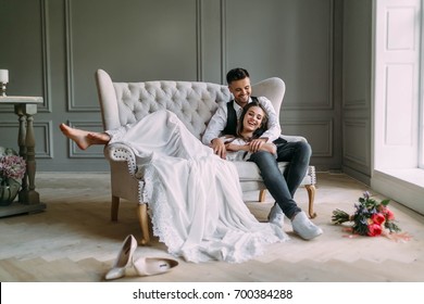Young bride in tender lace dress lies on knees of a handsome groom in blue jeans. Cheerful newlyweds are laughing. Full length. Indoors