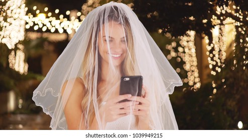 Young bride in wedding dress and veil taking a selfie with cell phone outdoors - Powered by Shutterstock