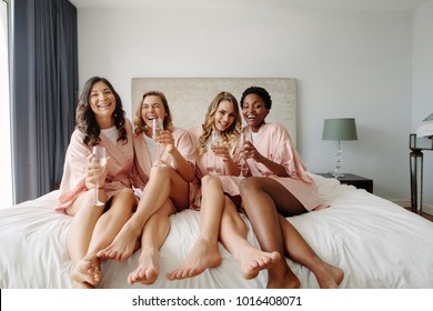 Young Bridesmaids In Silk Robes Drink Champagne In Hotel Room. Beautiful Females Celebrating Bachelorette Party, Sitting On Bed And Having Champagne.