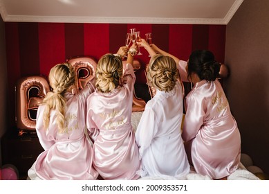 Young bridesmaids in silk robes drink champagne in hotel room. Beautiful females celebrating bachelorette party, sitting on bed. Back view.