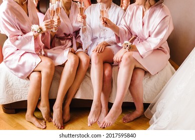 Young bridesmaids in silk robes drink champagne in hotel room. Beautiful females celebrating bachelorette party, sitting on bed.