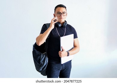 Young Brown Latino Male Dressed In Black With A Backpack, Glasses, Headphones, Holding A Laptop Computer, On A White Background, Generation Z, Student Concept Photograph.