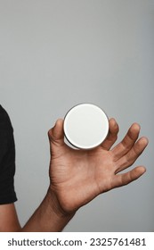 Young brown skin man holding up and showing in hand a round tin can packaging box for product, with white replaceable label. Mockup template, isolated from grey studio background.