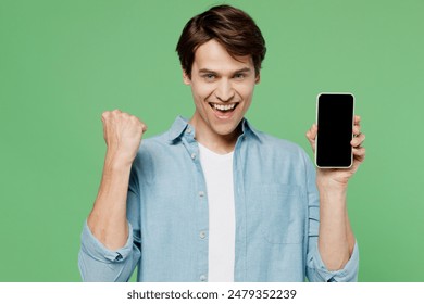 Young brunet man 20s wears blue shirt hold in hand use mobile cell phone with blank screen workspace area do winner gesture clenching fists say yes isolated on plain green background studio portrait - Powered by Shutterstock