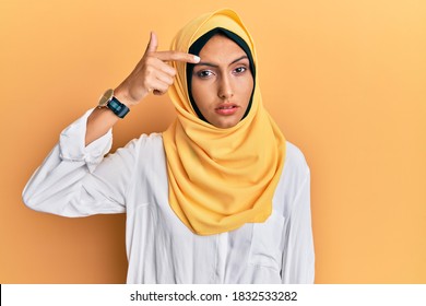 Young Brunette Arab Woman Wearing Traditional Islamic Hijab Scarf Pointing Unhappy To Pimple On Forehead, Ugly Infection Of Blackhead. Acne And Skin Problem 