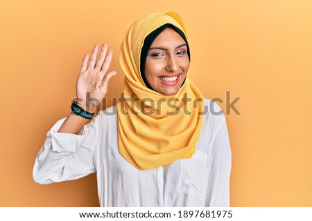 Young brunette arab woman wearing traditional islamic hijab scarf waiving saying hello happy and smiling, friendly welcome gesture 