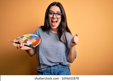 Young brunette artist woman painting holding painter brush and palette over yellow background screaming proud and celebrating victory and success very excited, cheering emotion