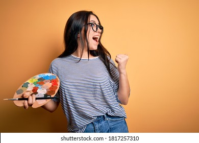 Young brunette artist woman painting holding painter brush and palette over yellow background pointing and showing with thumb up to the side with happy face smiling