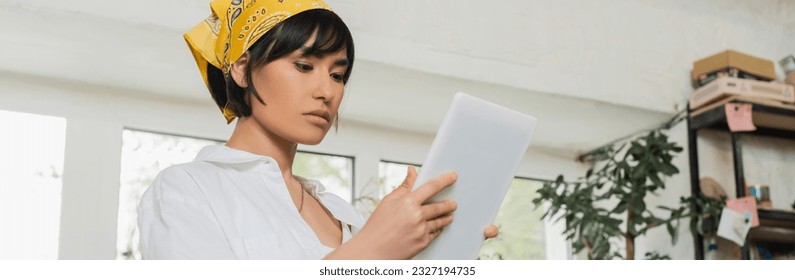 Young brunette asian artisan in headscarf and workwear using digital tablet while working in blurred ceramic workshop at background, craftsmanship in pottery making, banner - Powered by Shutterstock