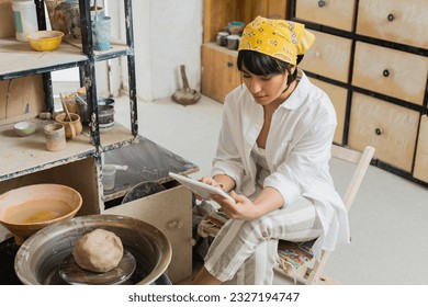 Young brunette asian artisan in headscarf and workwear using digital tablet while sitting near pottery wheel in blurred ceramic workshop, craftsmanship in pottery making - Powered by Shutterstock