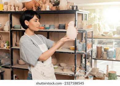 Young brunette asian female artisan in apron holding clay sculpture near rack with shelves in blurred ceramic workshop at background at sunset, artisan creating unique pottery pieces