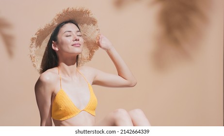 Young Brunette Asian Woman In Straw Hat And Yellow Crochet Bikini Enjoys The Sun, Sunbathing And Smiles For The Camera | Sunscreen Protection And Skin Care Commercial Concept