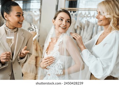 A young brunette bride in a wedding dress excitedly smiles while her middle-aged mother and best friend, a bridesmaid, help her put on a veil. - Powered by Shutterstock