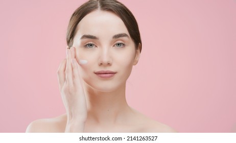 Young Brunette Caucasian Woman Applies Face Care Cream On Her Cheek On Pink Background | Face Cream Commercial Concept